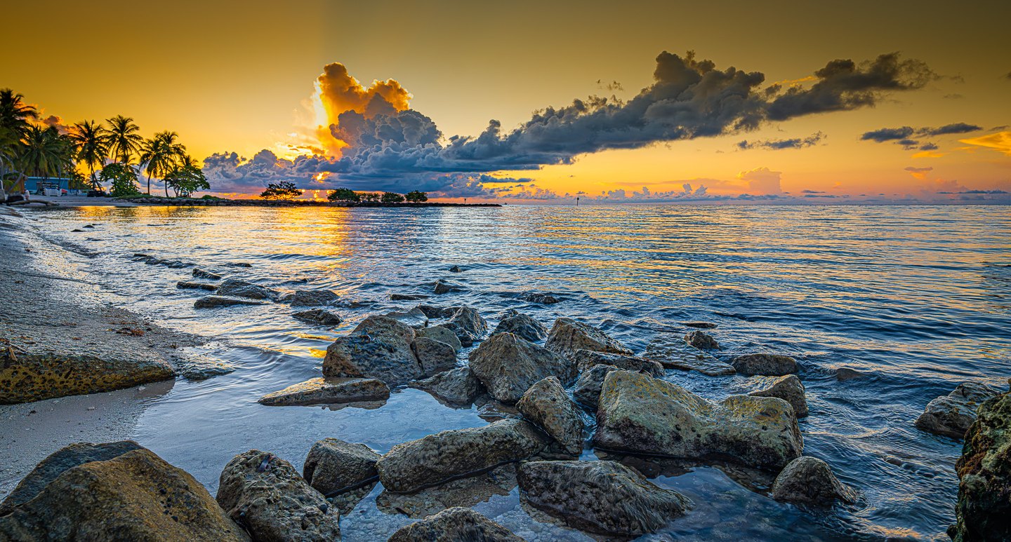 Waves wash around foreground rocks beneath a golden sky, with dark clouds and palms along the left shore.