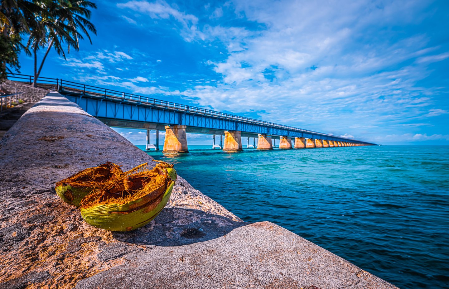 A split coconut husk rests on a stone seawall beside turquoise water and a long bridge beneath a cloud-streaked sky.