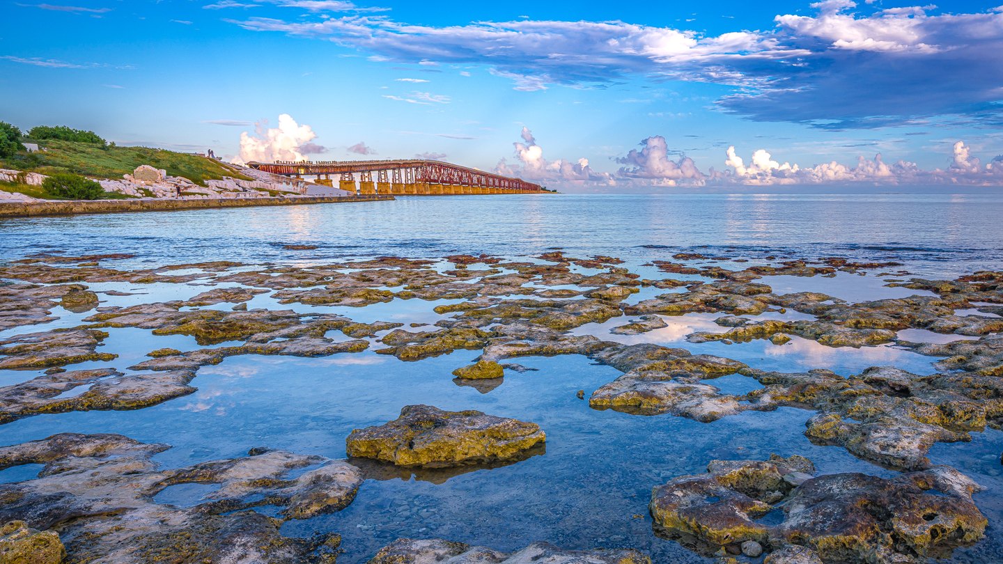 Rock pools reflect blue sky beside a calm sea, with a rust-colored bridge extending from a green shoreline.
