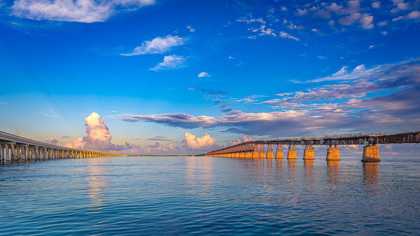 Two long bridges converge toward a distant shore over rippling blue water beneath clouds lit by golden sunlight.