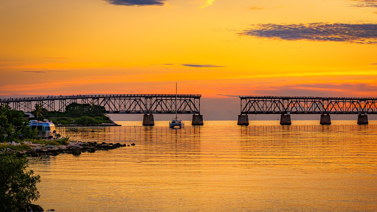 A sailboat rests before a bridge with a missing span above golden water, with a silver trailer on the rocky shore.