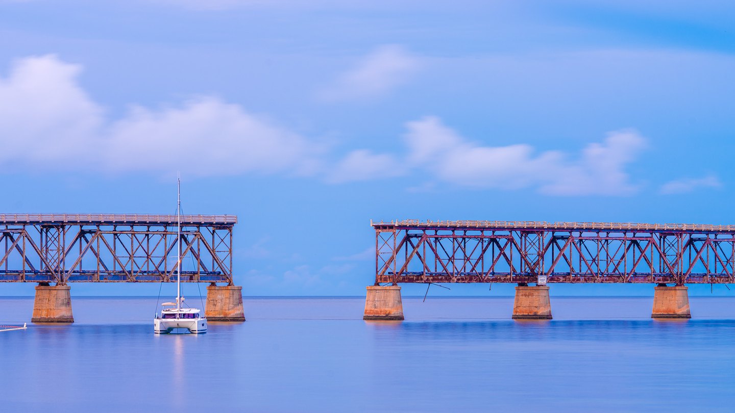 A white catamaran floats before two separated steel bridge spans on concrete piers beneath a blue, clouded sky.