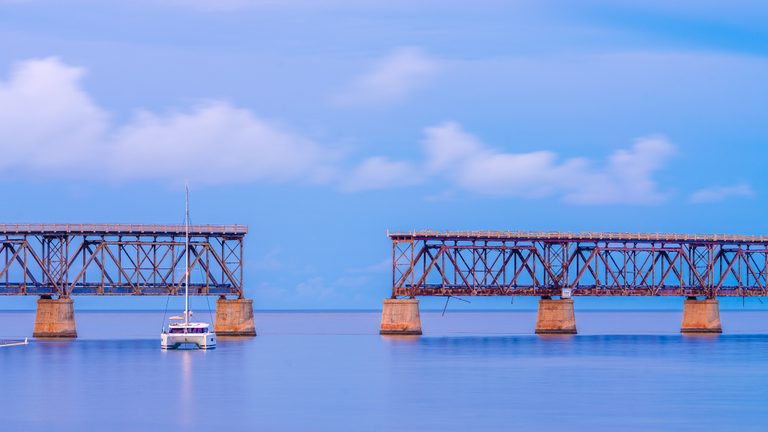 A white catamaran floats before two separated steel bridge spans on concrete piers beneath a blue, clouded sky.