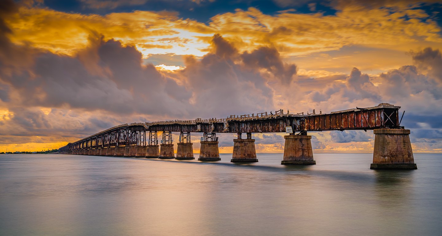 A rusted steel bridge on concrete piers stretches across calm water beneath towering gray and golden clouds.