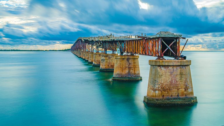 A rusted, broken bridge on concrete piers recedes across turquoise water beneath sweeping blue-gray clouds.