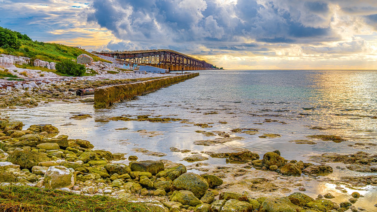 Rocky shore and a concrete seawall beside a long metal trestle, with golden light on the sea beneath large clouds.