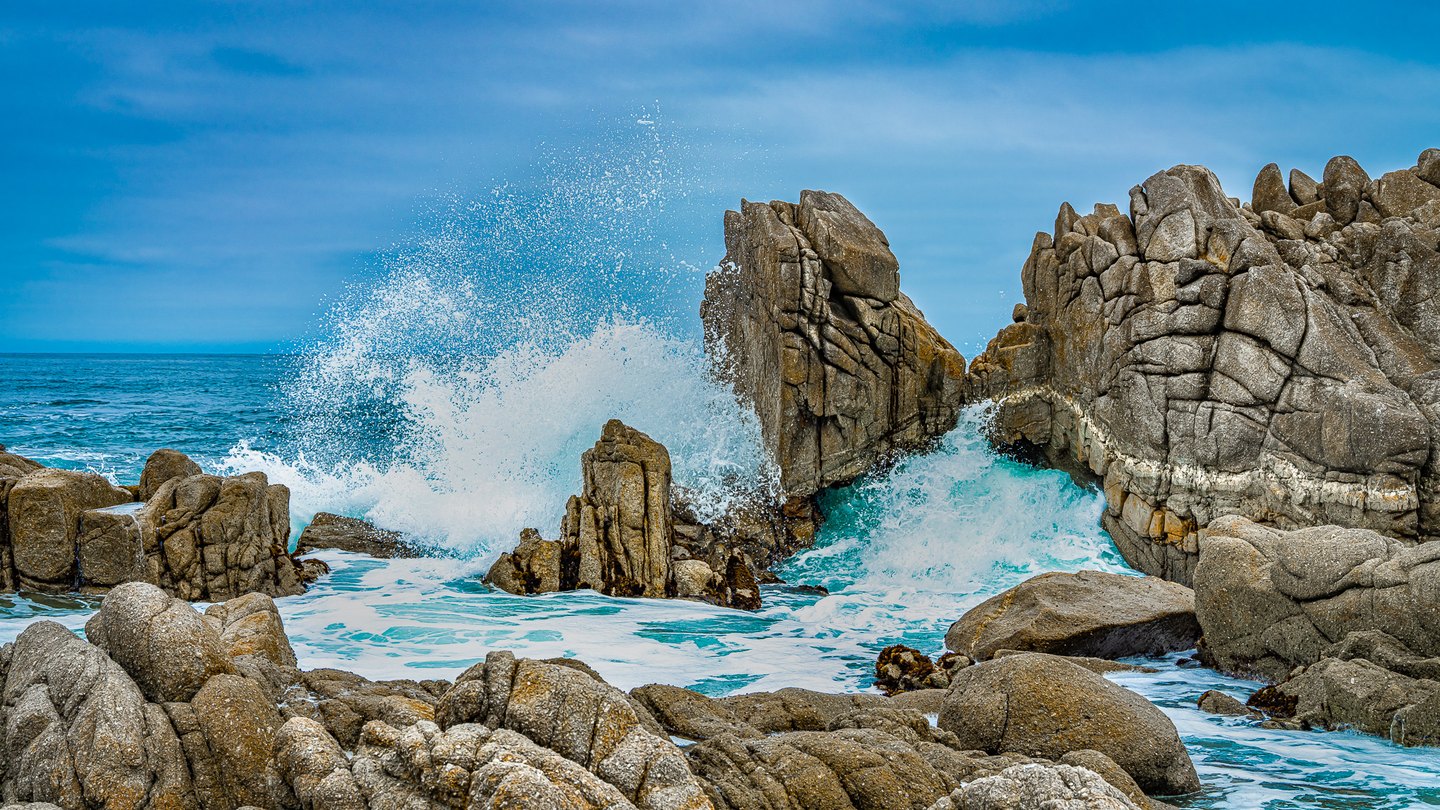 White surf bursts between jagged coastal rocks, with turquoise water below and a blue, lightly clouded sky above.