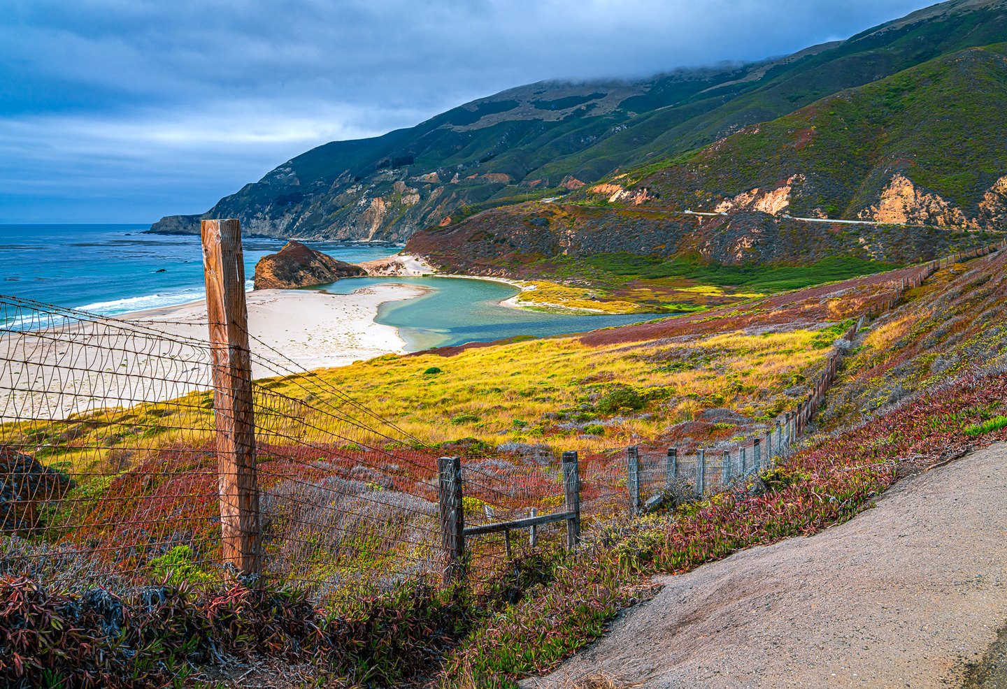 A wire fence crosses colorful coastal slopes above a turquoise lagoon, sandy beach, and ocean beneath clouded hills.