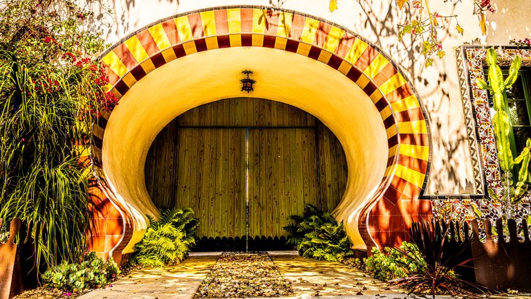 Wooden double doors sit within a yellow and terracotta tiled arch, surrounded by ferns, flowers, and a tall cactus.