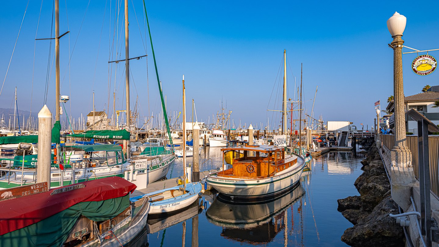 Sailboats and a varnished wooden motorboat reflect in calm Santa Barbara harbor water beside a waterfront walkway.