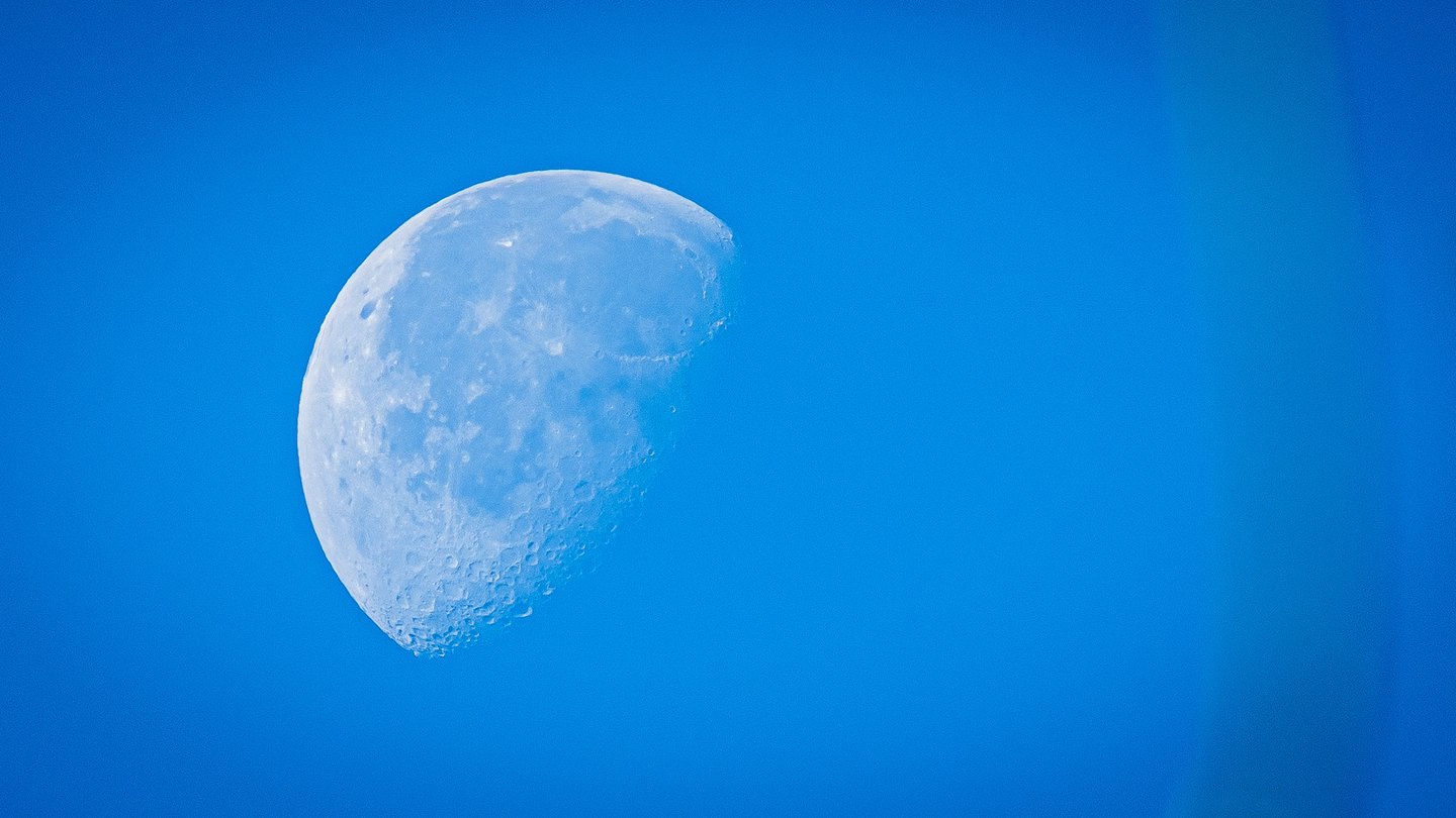 A pale, partially illuminated moon shows detailed craters against a clear blue sky, left of center.