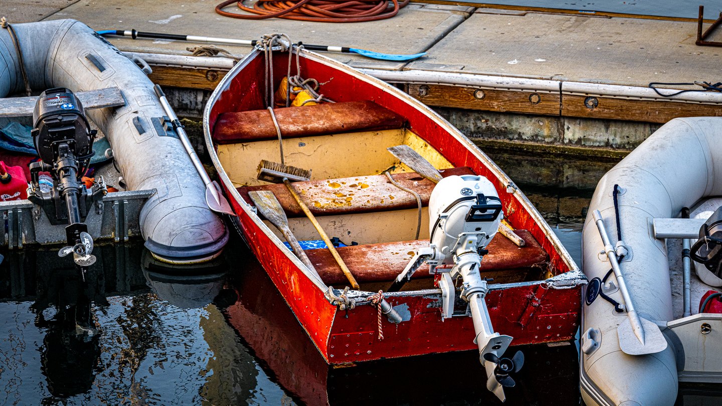 A weathered red dinghy with oars and an outboard motor floats between two gray inflatable boats beside a dock.