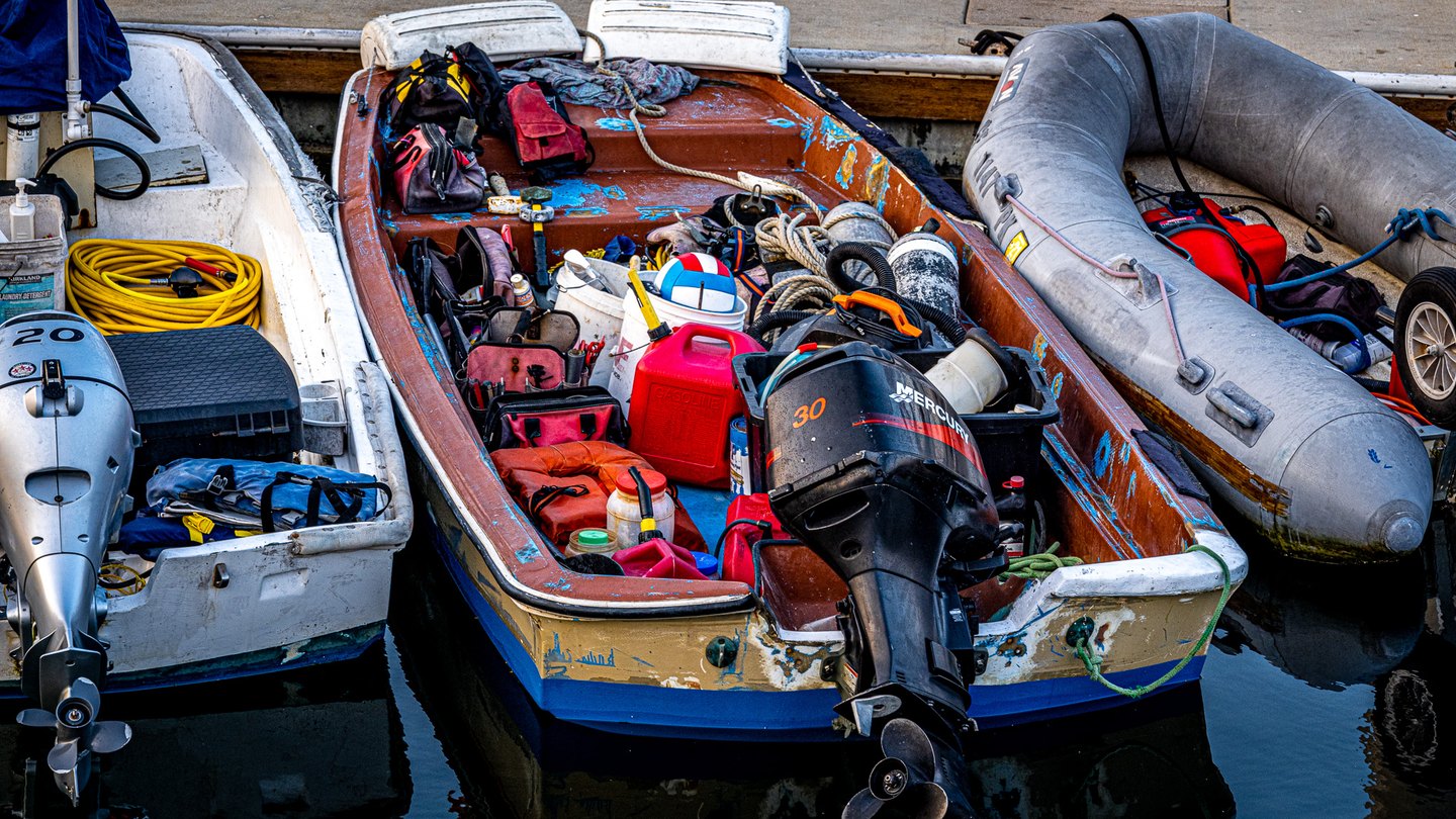 Three boats sit beside a dock, packed with ropes, fuel cans, tools, and bags, with outboard motors above dark water.