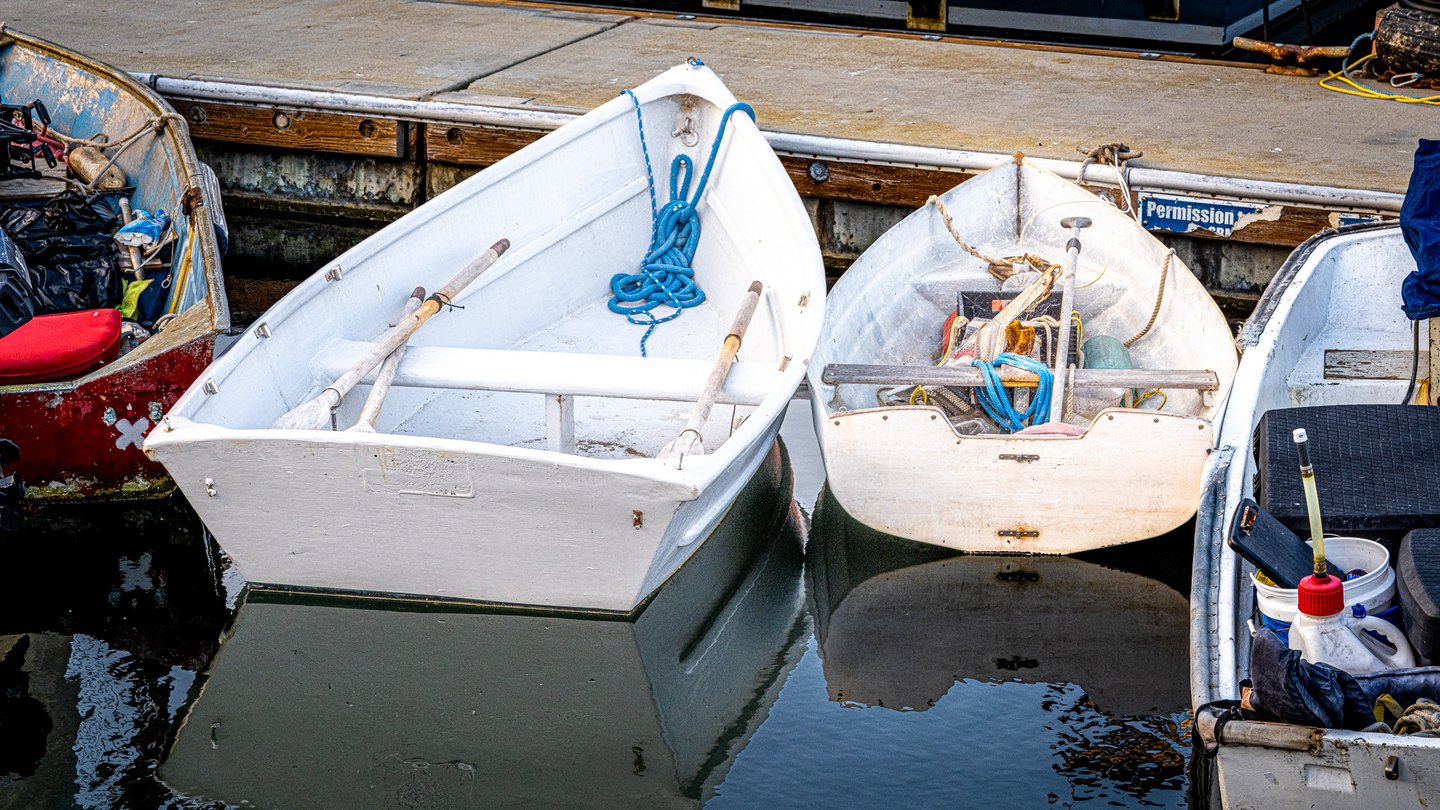 Small boats moored beside a dock hold oars, blue ropes and gear, their pale hulls reflected in dark water.