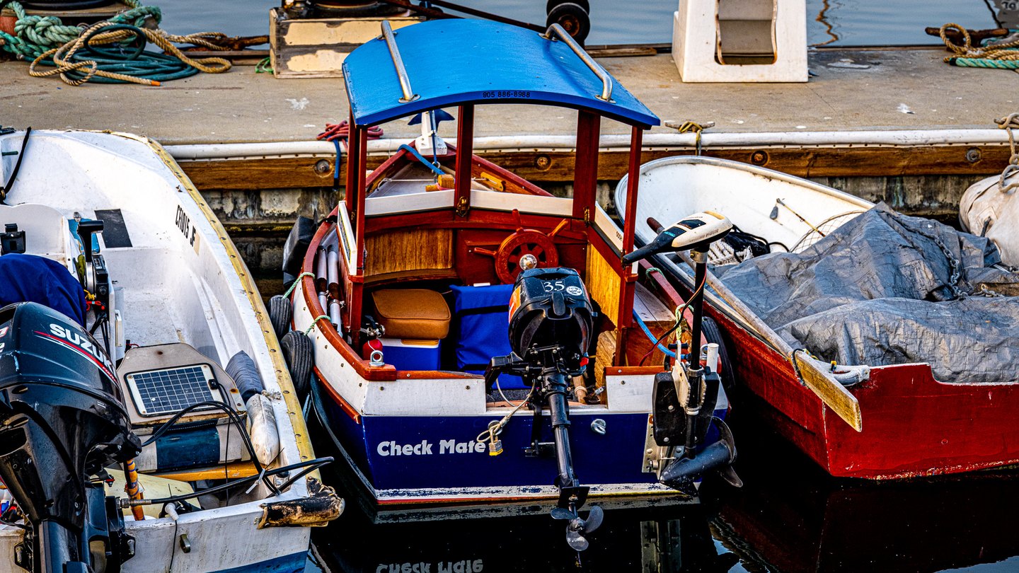 A wooden motorboat with a bright blue canopy sits between white and red boats beside a rope-strewn dock.