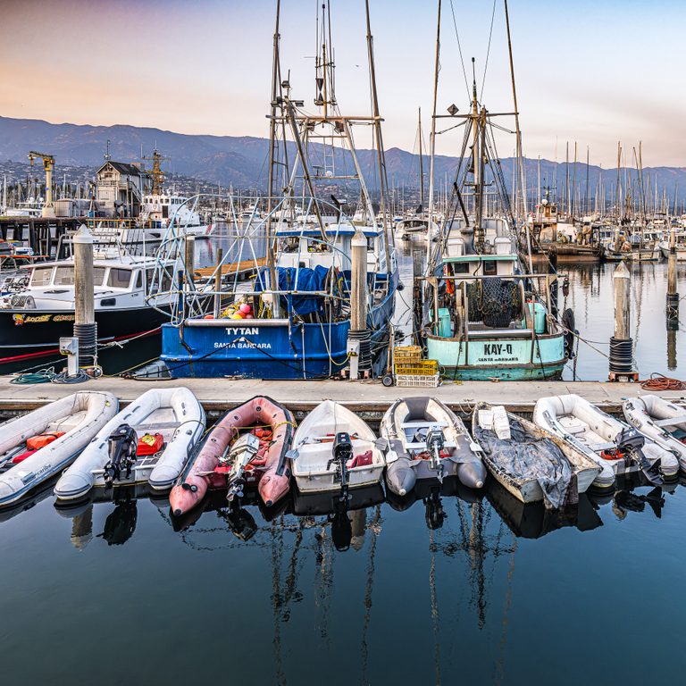 Dinghies and blue and turquoise fishing boats line a calm harbor, with tall masts and mountains behind them.