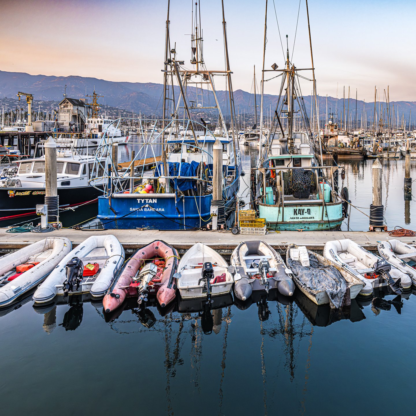 Dinghies and blue and turquoise fishing boats line a calm harbor, with tall masts and mountains behind them.
