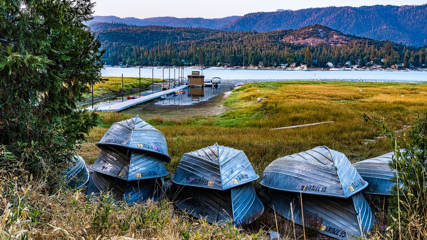 Upturned metal boats sit beside golden grasses, with a dock, blue lake, lakeside houses, and forested ridges beyond.