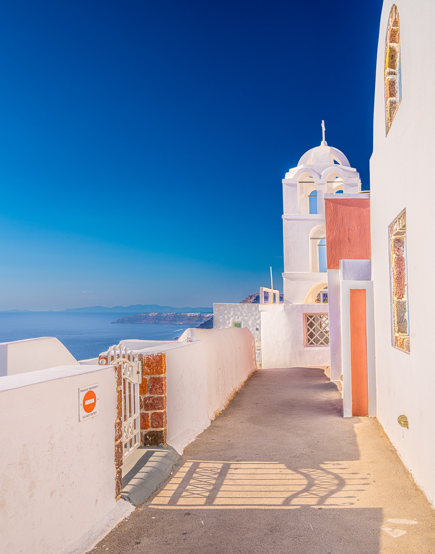A sunlit lane between white and coral walls leads toward a white church tower, with blue sea and distant cliffs beyond.