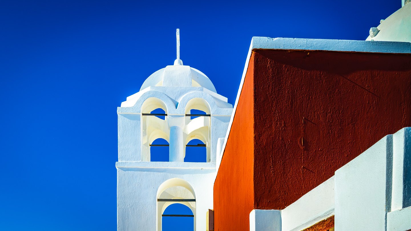 A white arched tower rises against a deep blue sky beside an orange-red wall edged in white.