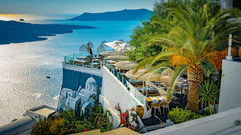 A seaside dining terrace with woven shades, a large palm and white walls overlooks sparkling blue water and dark islands.