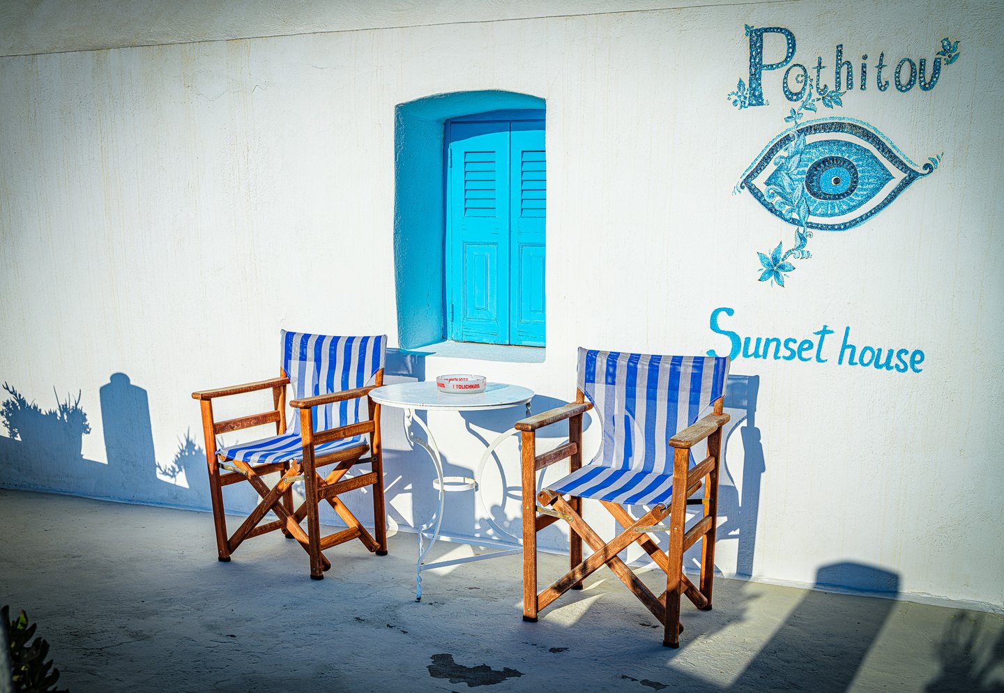 Two blue-and-white striped chairs flank a white table beneath turquoise shutters and a painted eye on a white wall.