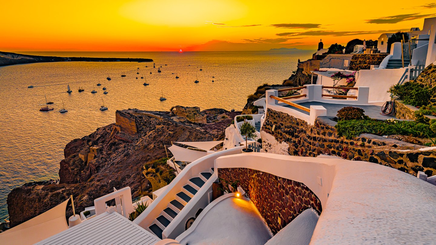 White cliffside terraces and winding stairs overlook sailboats on a golden sea beneath an orange sunset.