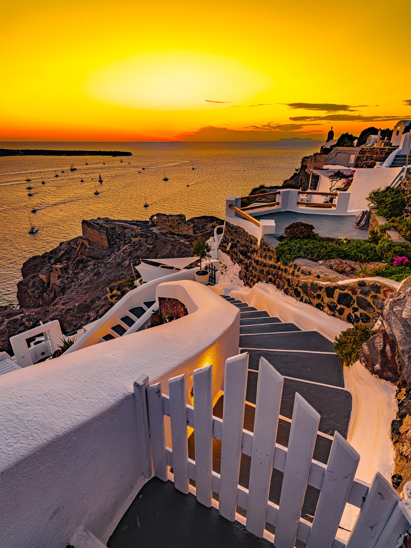 A white gate and winding cliffside stairs overlook sailboats on a golden sea beneath an orange sunset sky.