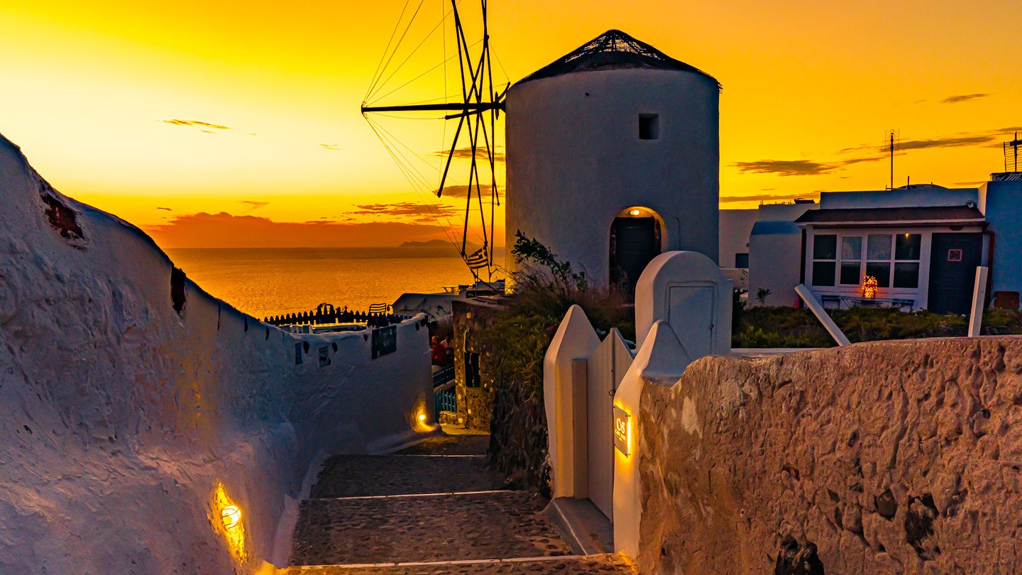 A stepped lane between whitewashed walls leads toward a windmill and the sea beneath a golden sunset.
