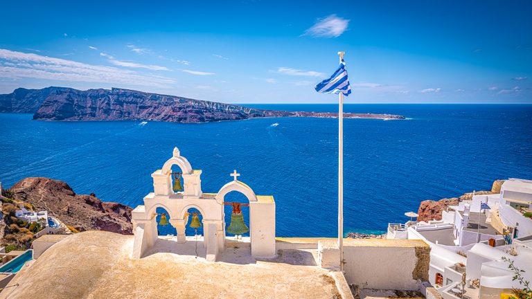 White bell arches and a Greek flag overlook a deep blue sea, rocky cliffs, and whitewashed hillside buildings.
