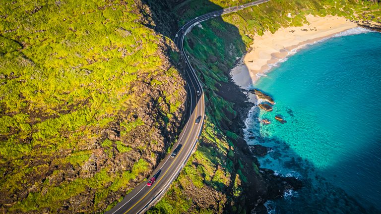 A coastal road curves between sunlit green cliffs and turquoise water, passing a sandy beach and dark shoreline rocks.