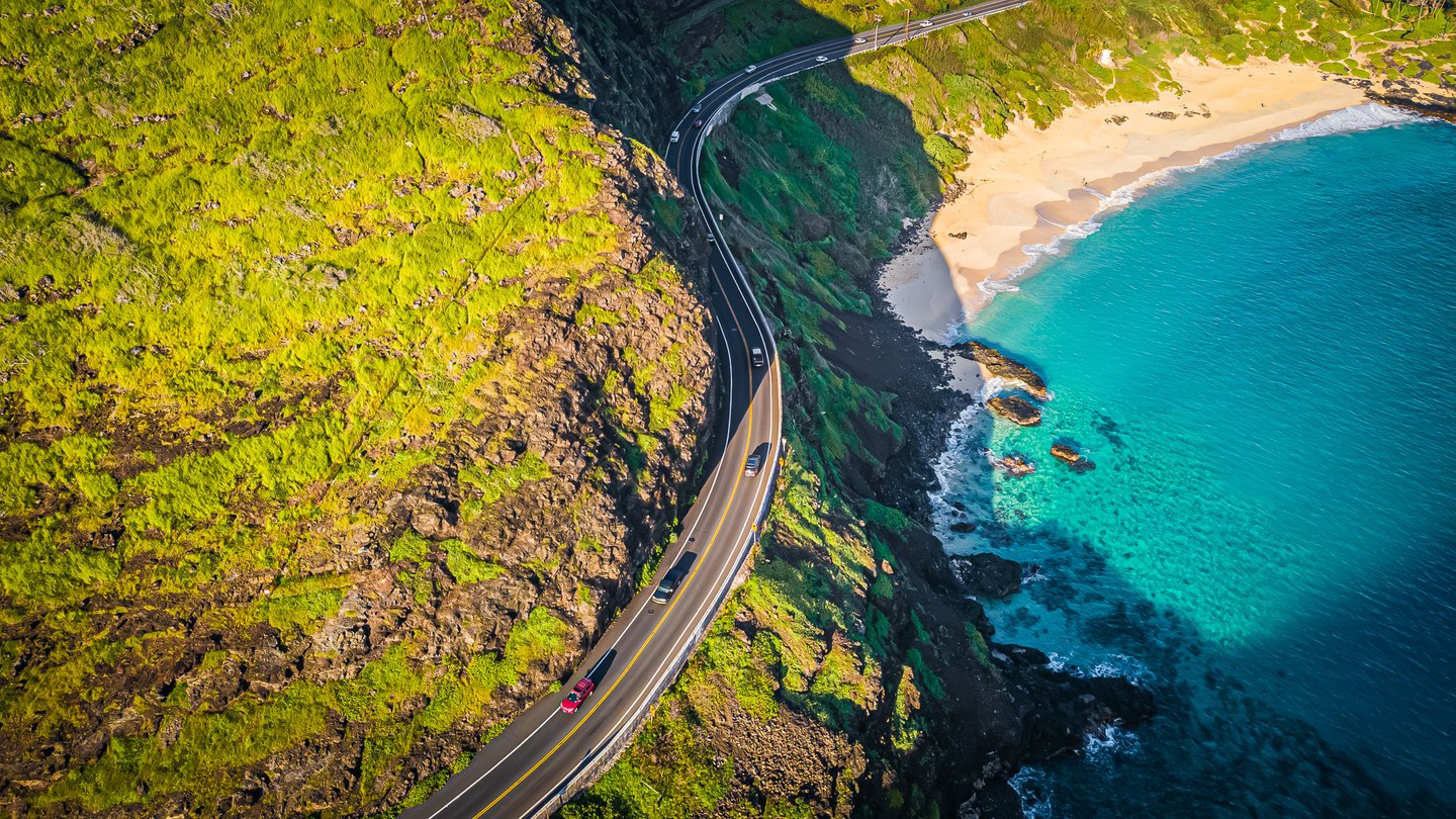 A coastal road curves between sunlit green cliffs and turquoise water, passing a sandy beach and dark shoreline rocks.